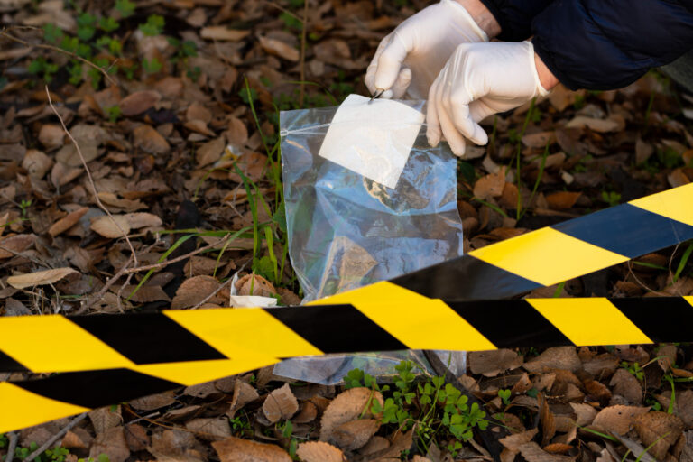 Crime scene tape with evidence being collect into bag by person wearing gloves.